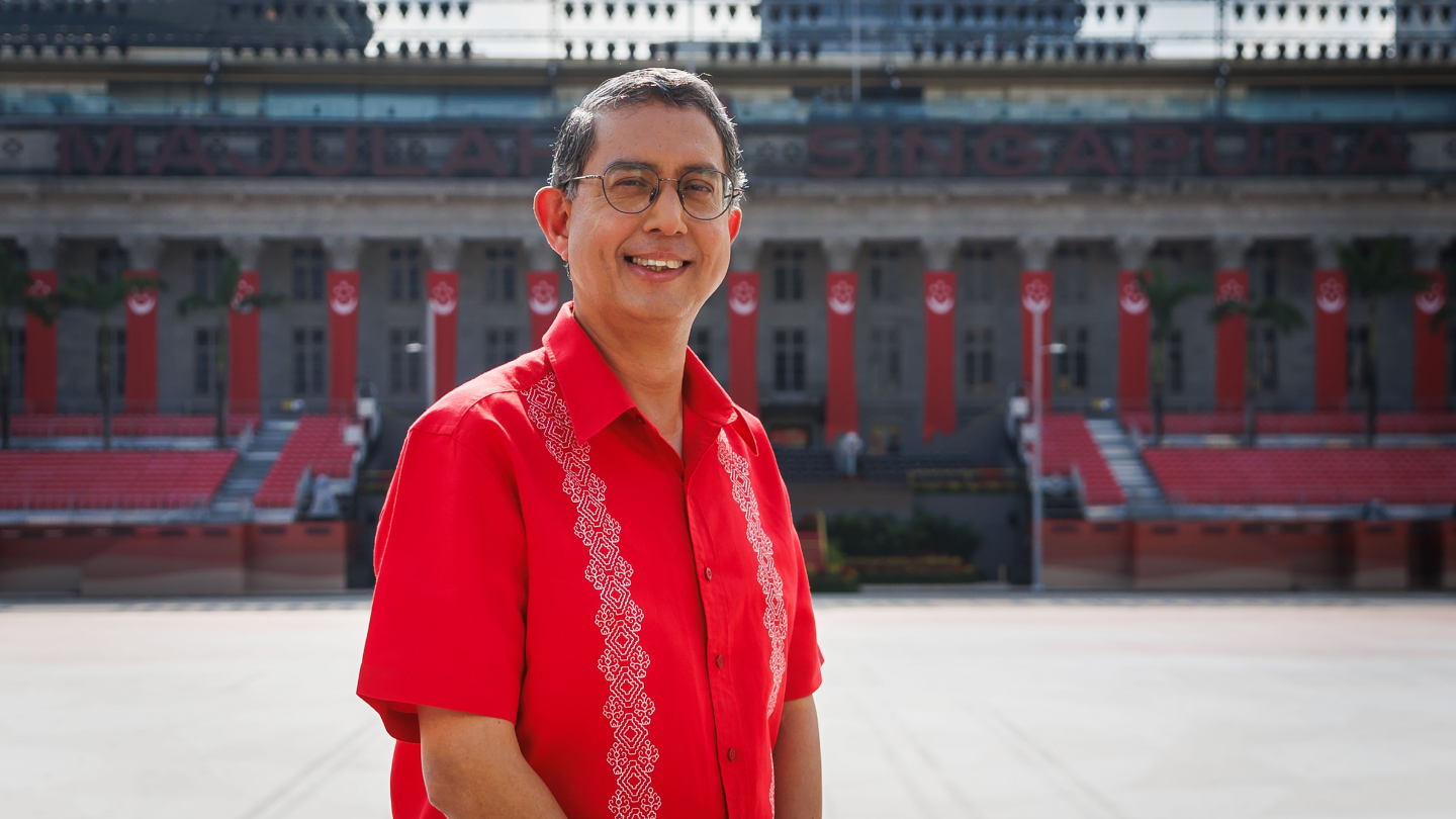Man in red shirt at civic square; grandstand and “Majulah Singapura” banners behind.